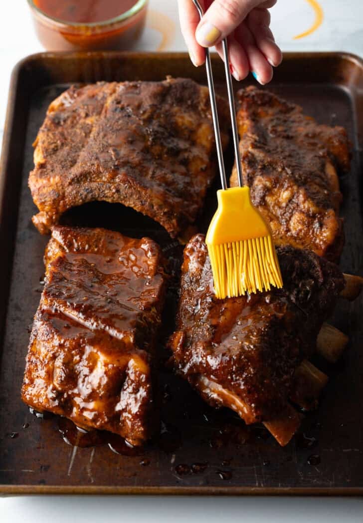 4 sections of cooked baby back ribs on a baking sheet. A yellow basting brush is brushing on bbq sauce.