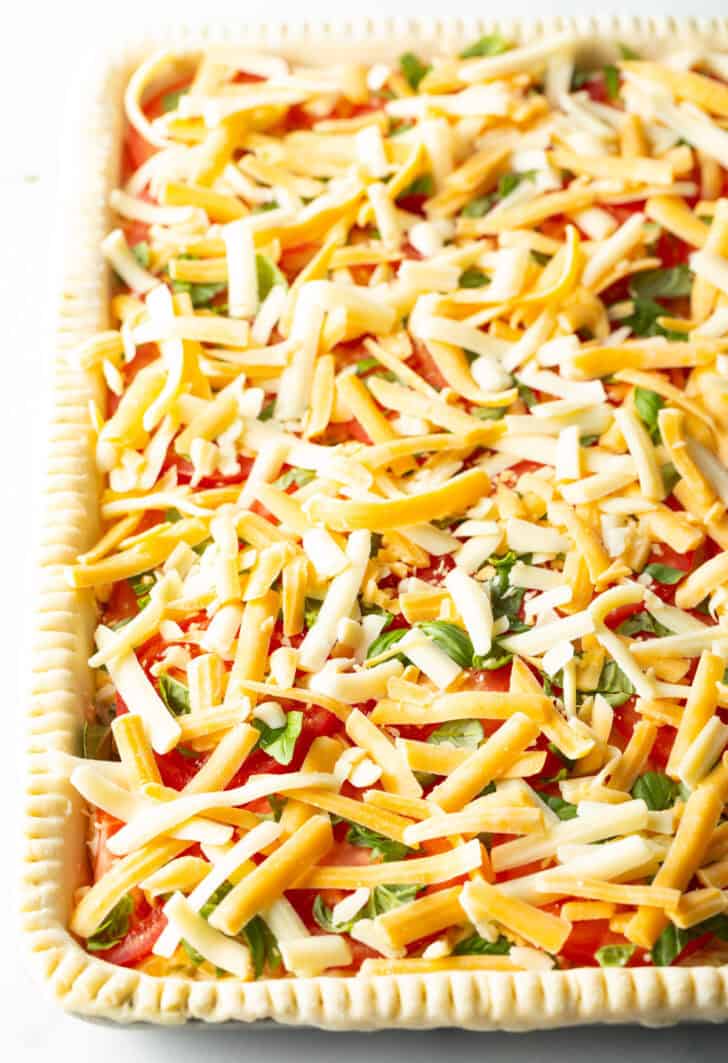 Top down view of raw slices of tomato in an unbaked slab pie, topped with shredded cheese and sliced green onions.