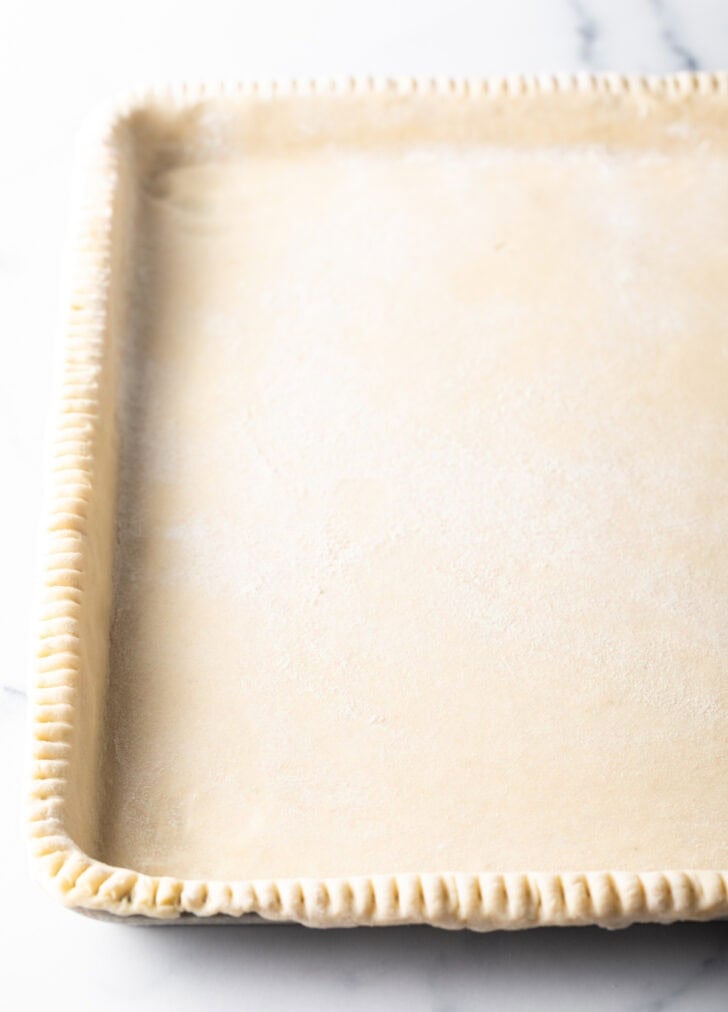 Raw pie dough crimped around a large rectangle baking sheet.