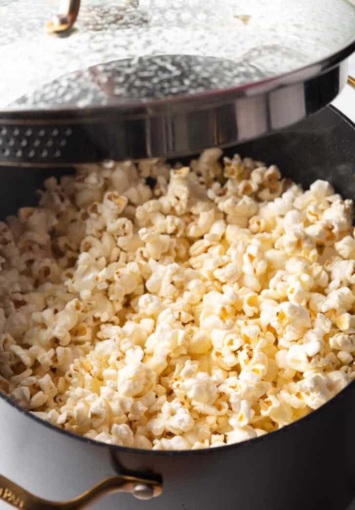 Opening the lid after cooking popcorn on the stovetop in a large black pot.