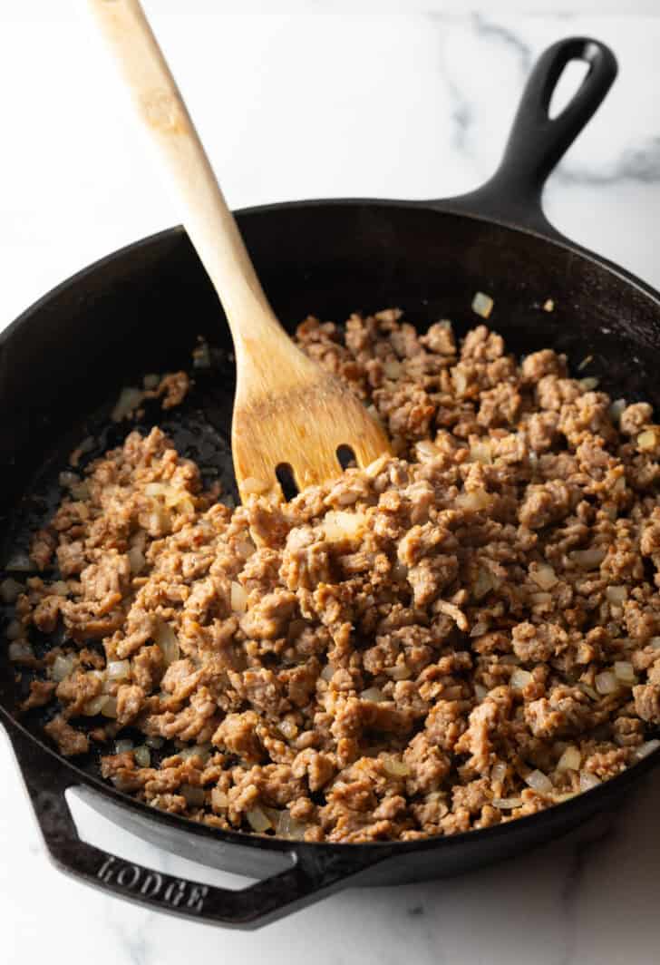 Browning ground beef in a large cast iron skillet with a wooden spatula.