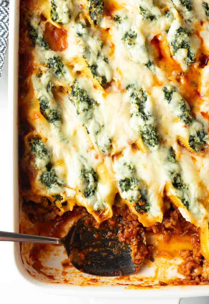 Top down view large rectangle baking dish with spinach and meat stuffed shells, with a serving taken out.