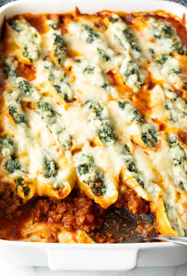 Top down view large rectangle baking dish with spinach and meat stuffed shells, with a serving taken out.