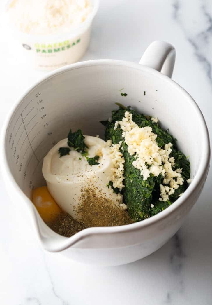 Large white mixing pitcher with ricotta cheese, cooked spinach, garlic and egg.