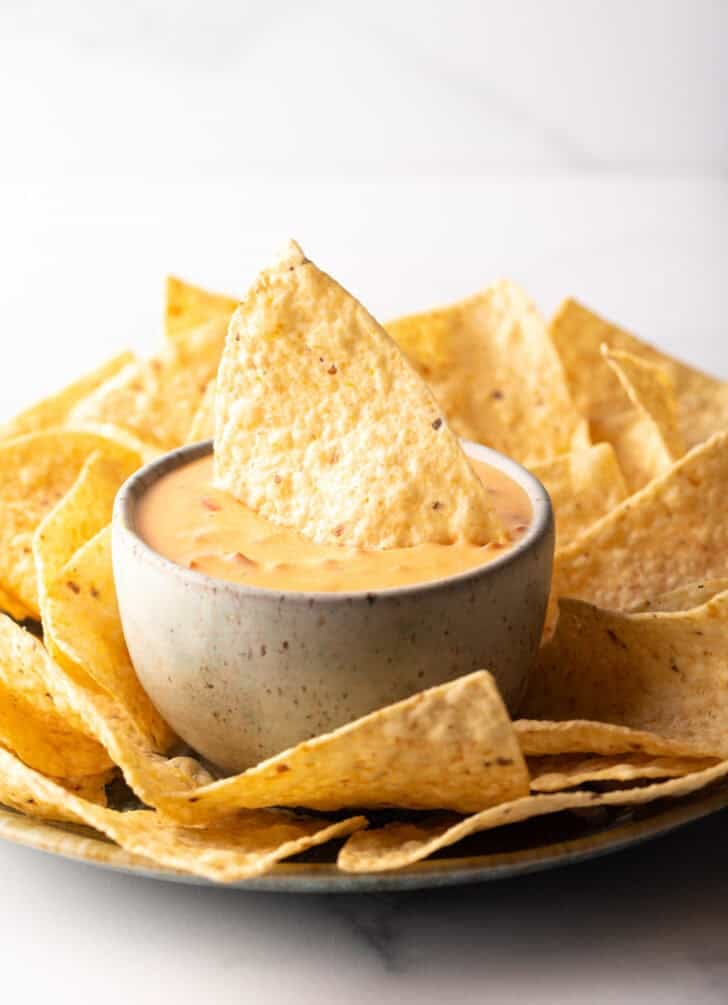 Chip in a white bowl of velveeta cheese dip surrounded by tortilla chips.