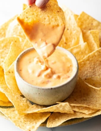 Hand dipping a chip into a white bowl of velveeta cheese dip surrounded by tortilla chips.