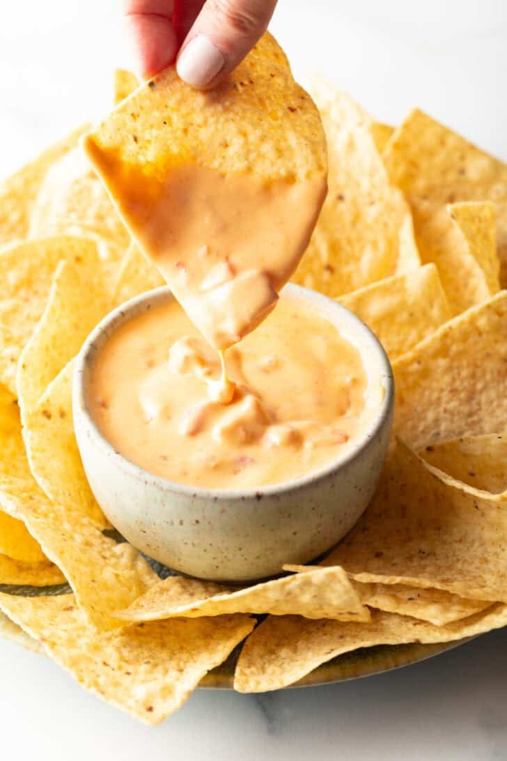 Hand dipping a chip into a white bowl of velveeta cheese dip surrounded by tortilla chips.