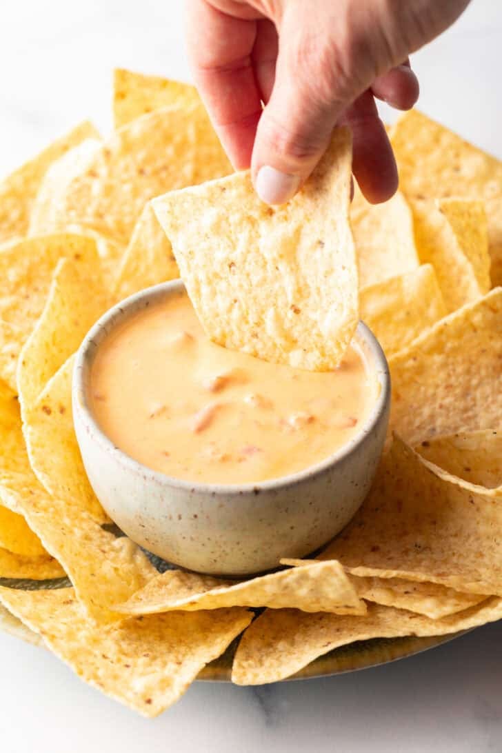 Hand dipping a chip into a white bowl of velveeta cheese dip surrounded by tortilla chips.