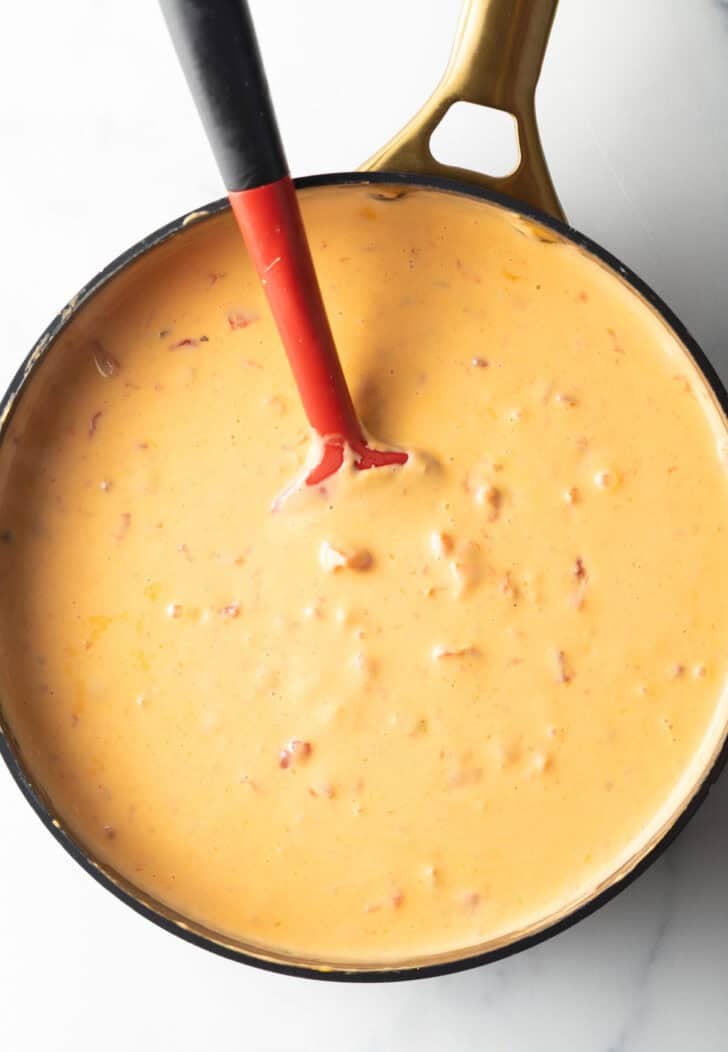 Using a red spatula to stir velveeta cheese and rotel dip in a deep black skillet.