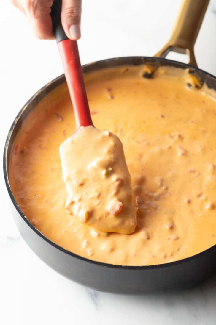 Using a red spatula to stir velveeta cheese and rotel dip in a deep black skillet.