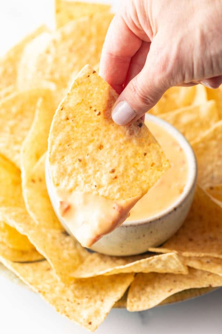 Hand holding a tortilla chip dipping in cheese dip to camera.