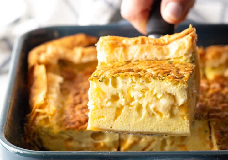 Hand removing a square of egg casserole with a spatula from a square baking dish.