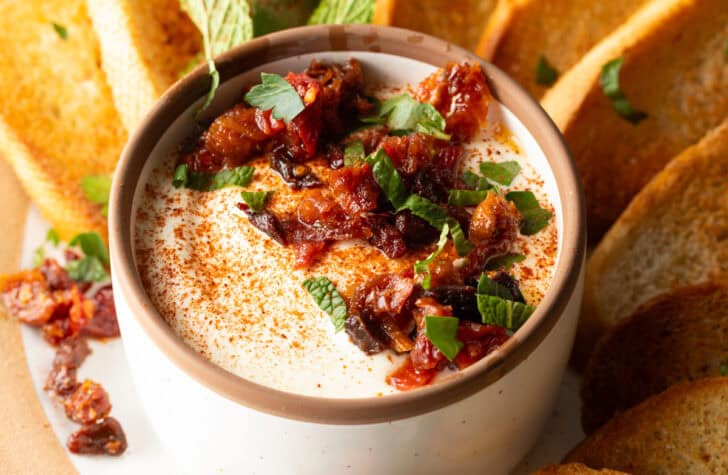 White cup with whipped feta with chopped sun dried tomato, on a plate surrounded by slices of toasted baguette.