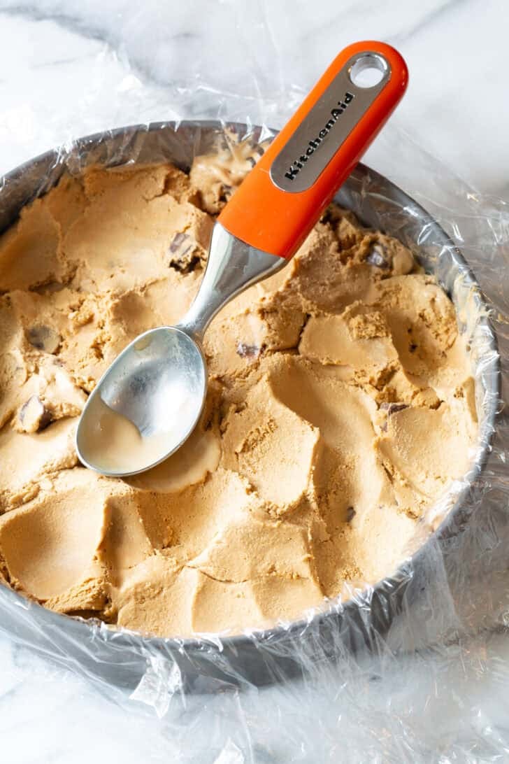 Using a metal scoop to smooth ice cream into a cake pan lined with plastic wrap, to create a thick layer of ice cream.