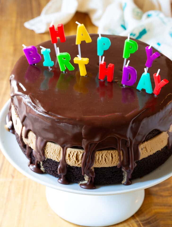 Chocolate and caramel ice cream layered cream cake coated with glossy icing and Happy Birthday candles on top.