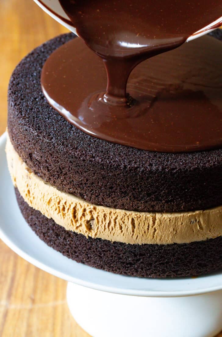 Pouring melted fudge frosting overtop a chocolate and caramel layered ice cream cake.