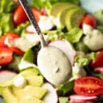 A spoon is drizzling homemade avocado lime ranch dressing over a salad with tomatoes and avocados.