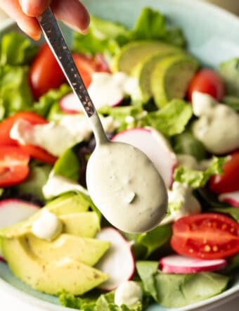 A spoon is drizzling homemade avocado lime ranch dressing over a salad with tomatoes and avocados.