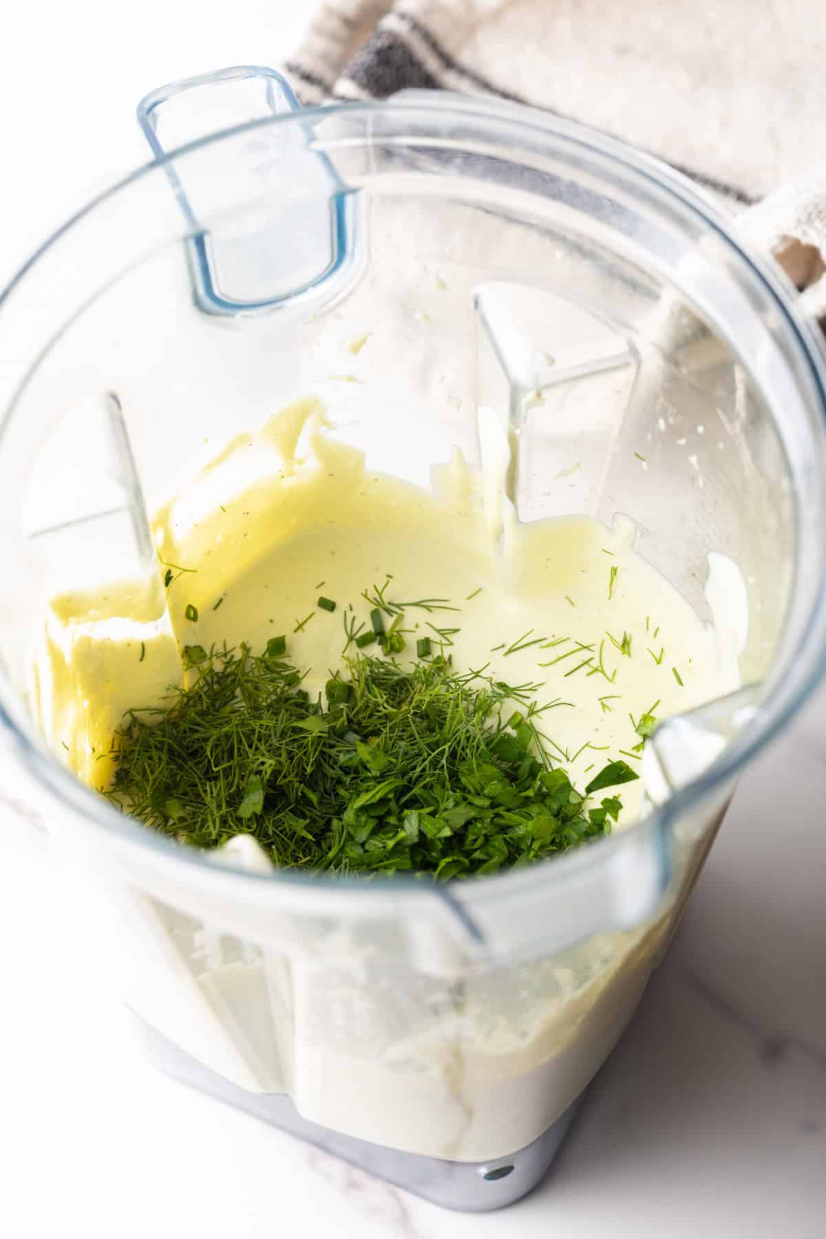 Fresh herbs are being incorporated into an avocado ranch dressing recipe with a blender.