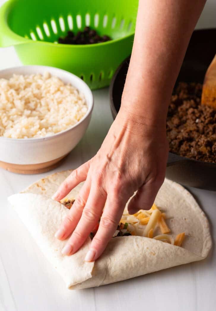 Hand folding a tortilla to make a burrito filled with beef and cheese.