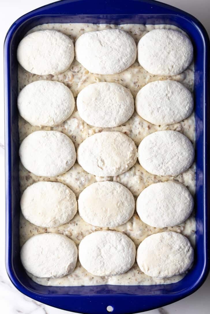 Frozen biscuits lined up on top of a blue pan filled with sausage gravy.
