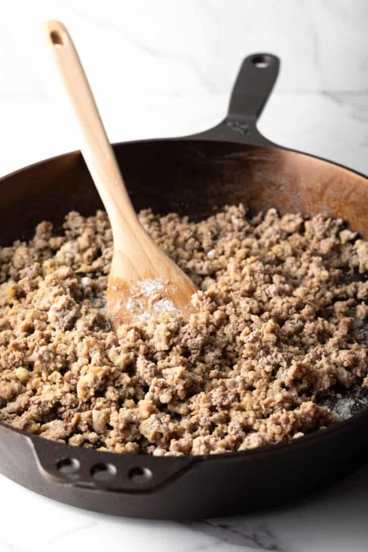 A black pan with cooked crumbled sausage being stirred with a wooden spoon.
