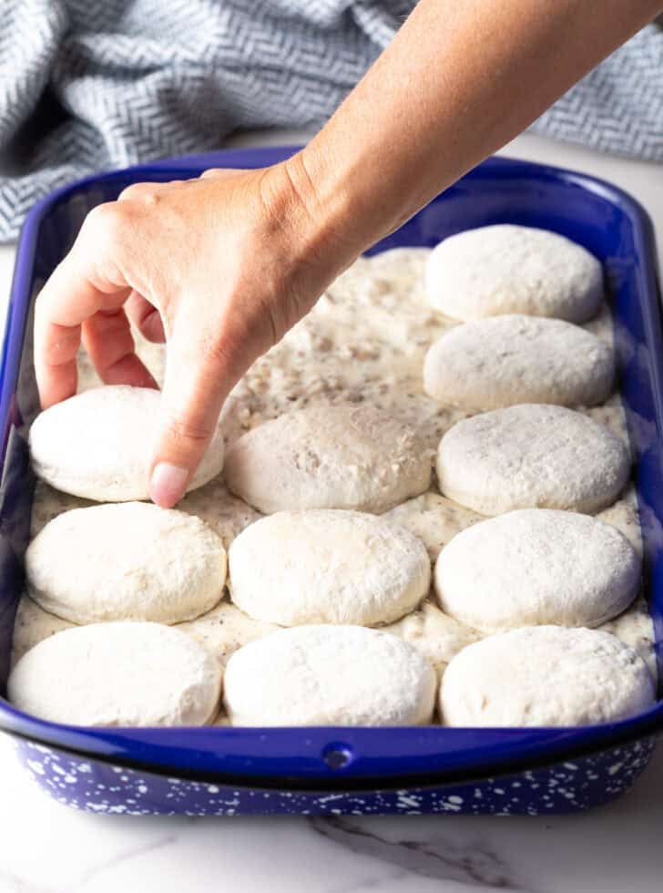 A hand arranging frozen biscuits on top of a blue pan filled with sausage gravy.