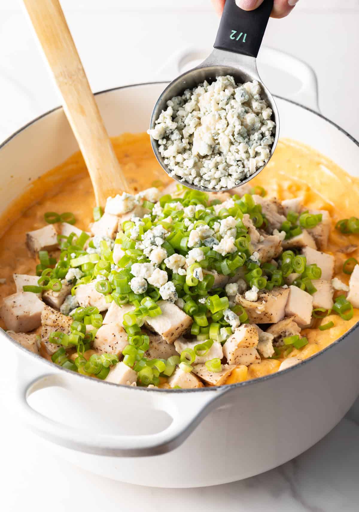 Adding crumbled blue cheese from a metal measuring cup onto a mound of cooked chopped chicken and chopped green scallions in a large pot of macaroni and cheese.