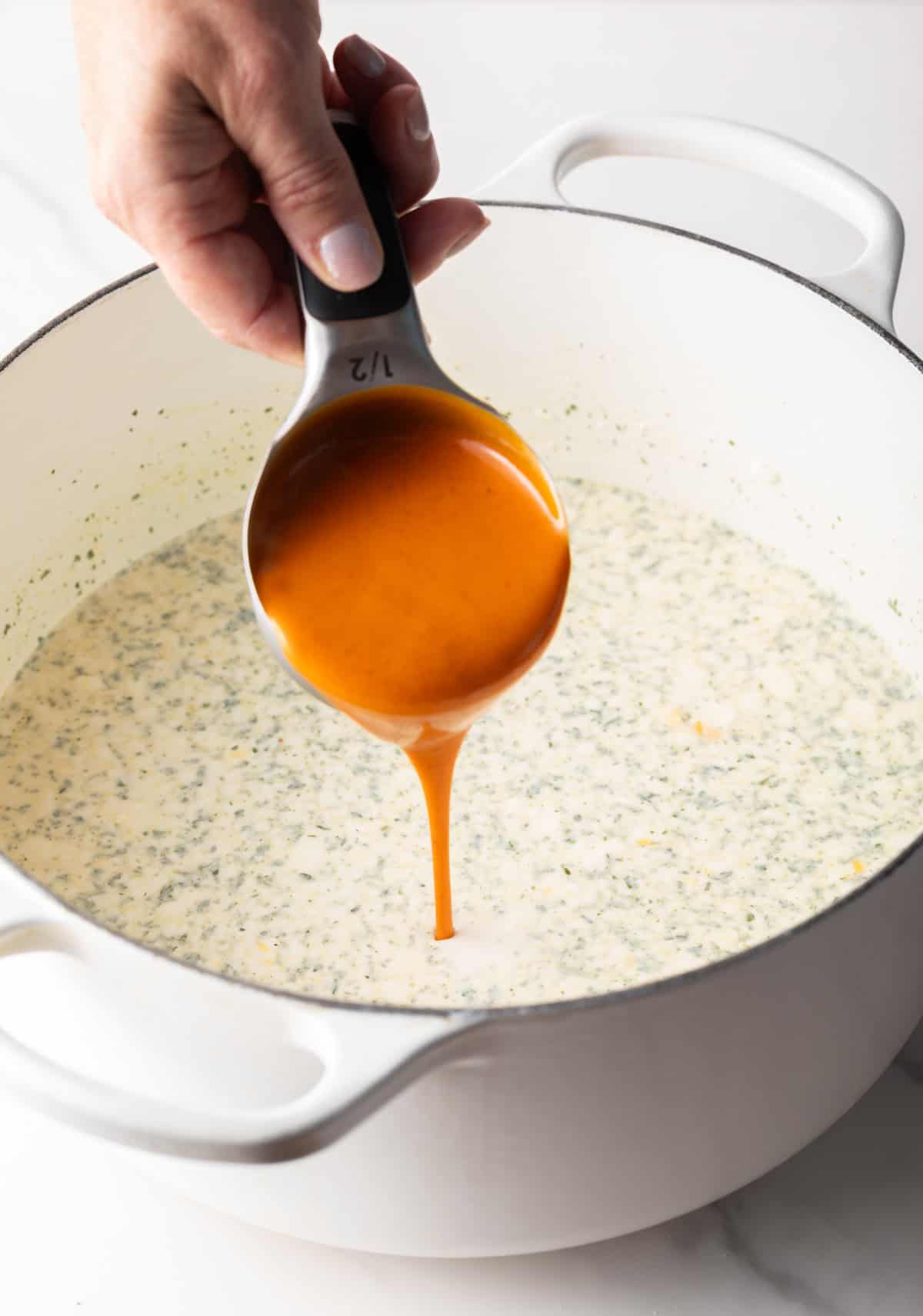 Hand pouring buffalo sauce from a metal measuring cup into a large white pot.