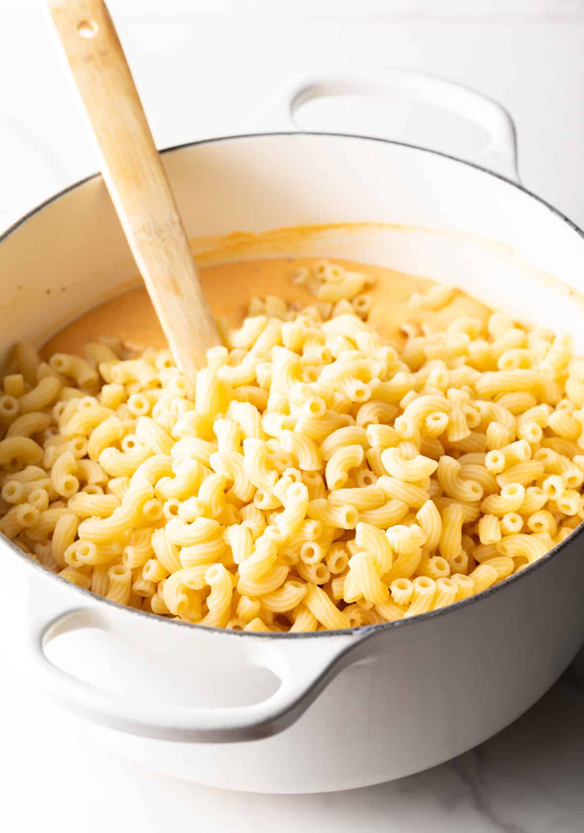 Stirring a large white pot of macaroni noodles with a wooden spatula.