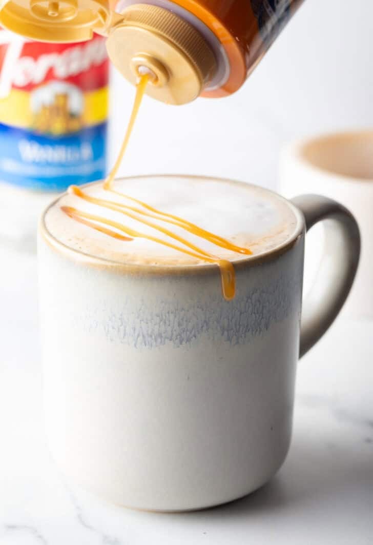 Drizzling caramel sauce from a bottle over a white mug with caramel macchiato.