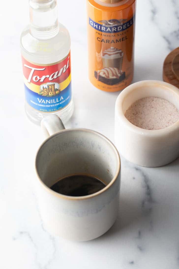 White mug of coffee on a marble background, with bottle of vanilla syrup and bottle of caramel.
