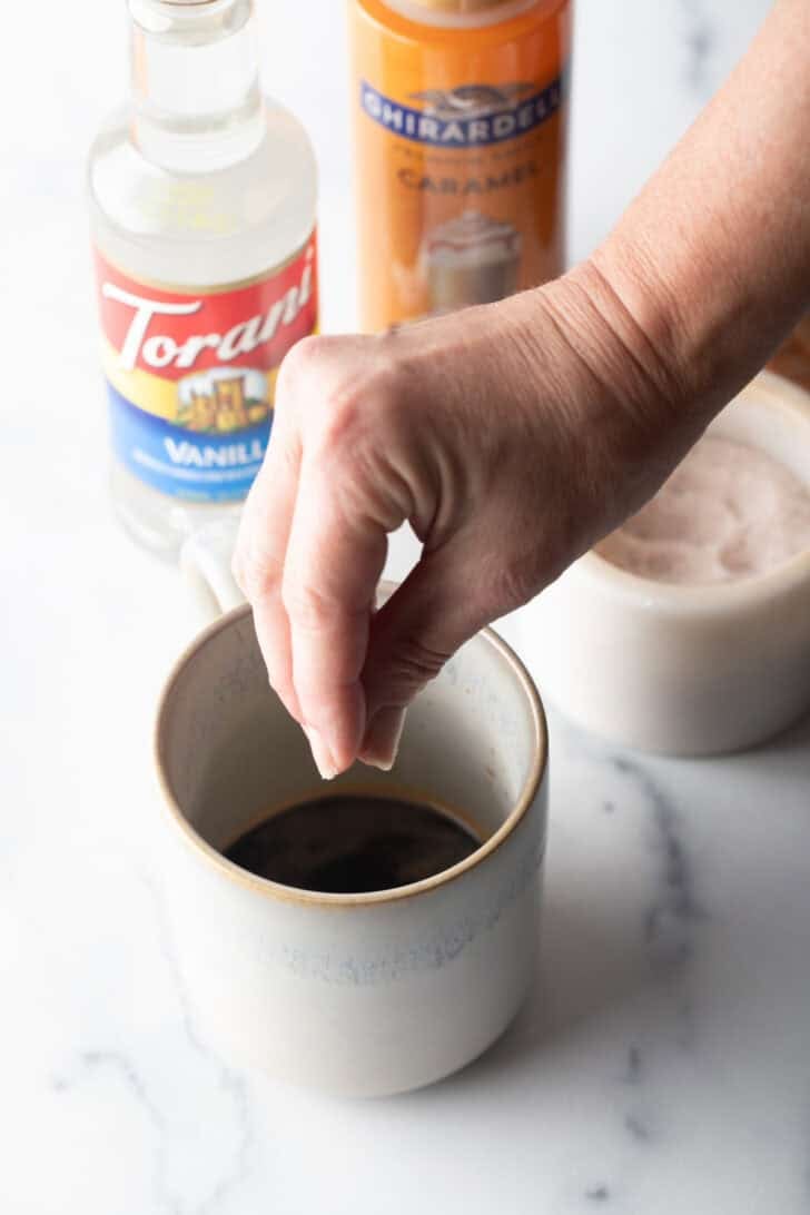Hand sprinkling salt into a white mug half filled with coffee.
