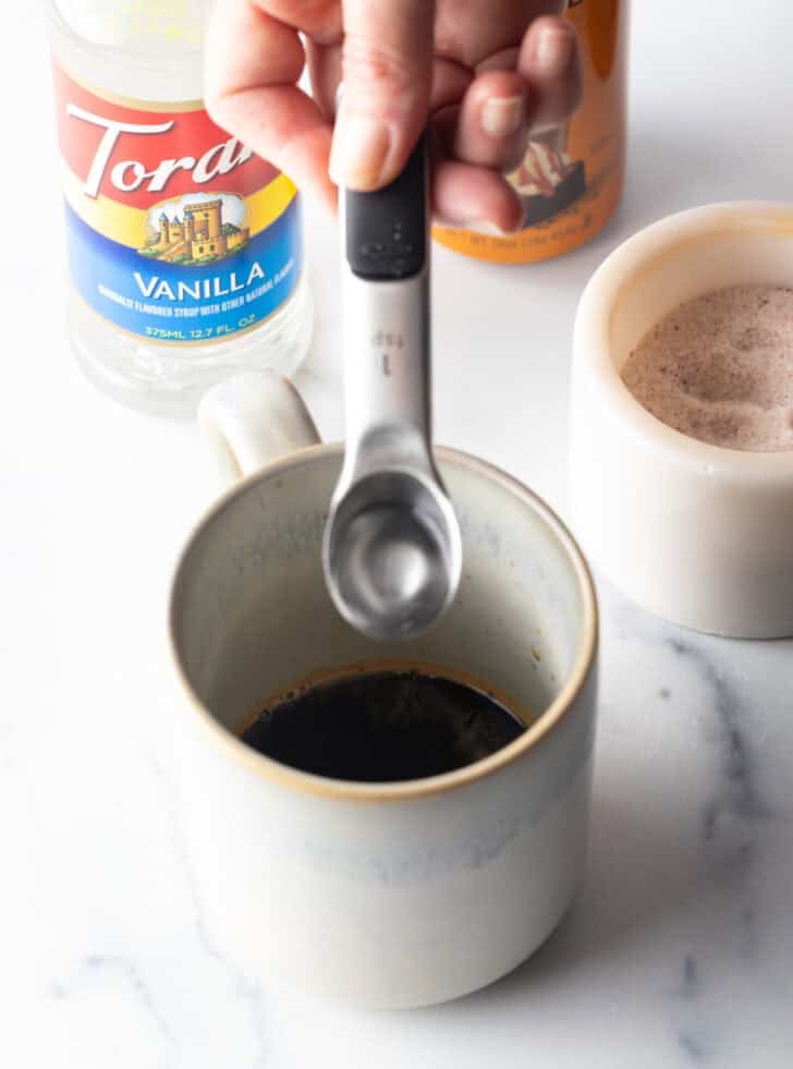 Hand adding vanilla syrup from a metal teaspoon into a white mug of coffee.