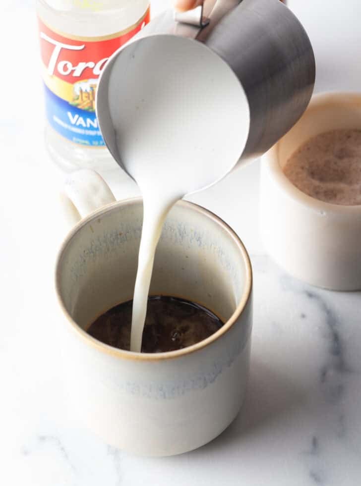 Pouring foaming milk from a metal pitcher into a white mug of espresso.