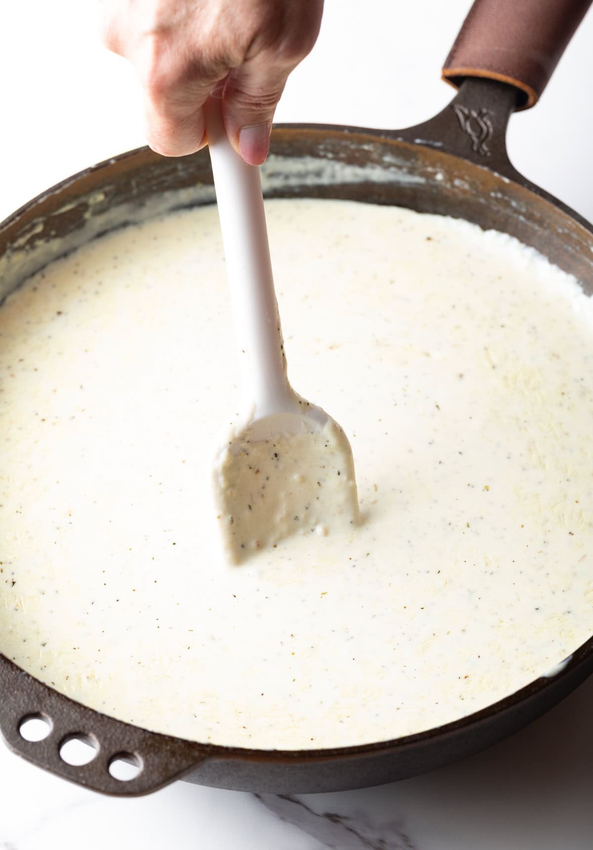 Hand with white spatula stirring alfredo sauce in a large skillet.