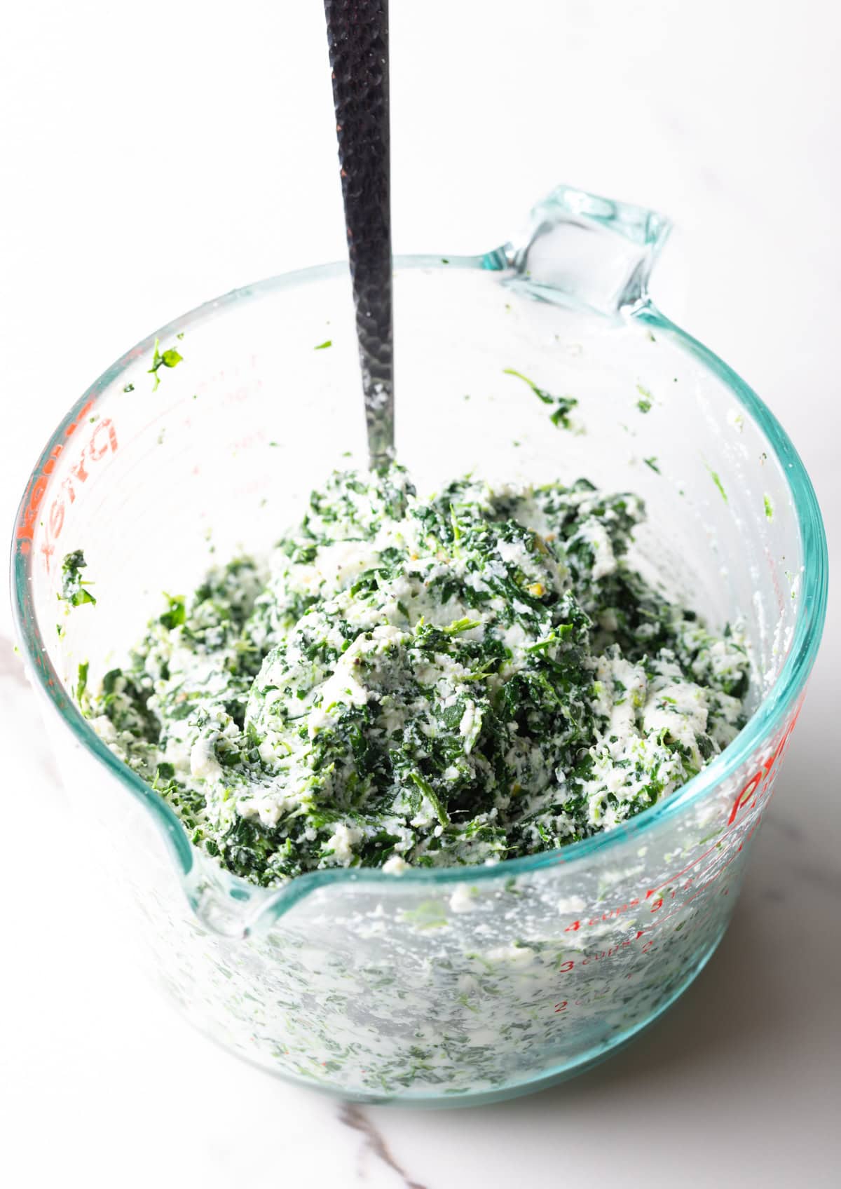Thawed frozen spinach and ricotta combined in a glass measuring cup with metal spoon.