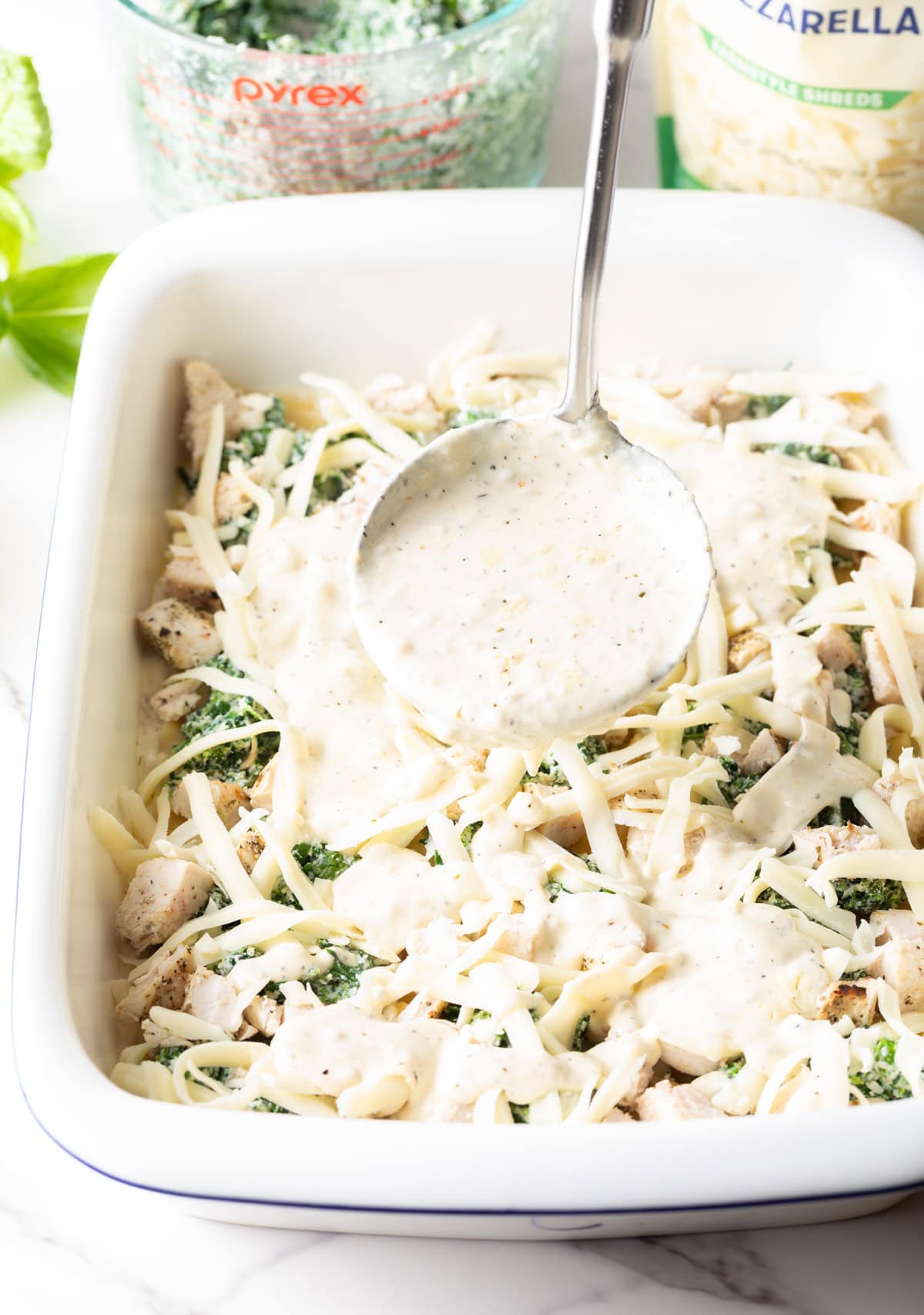 Using a ladle to pour alfredo sauce over the casserole.