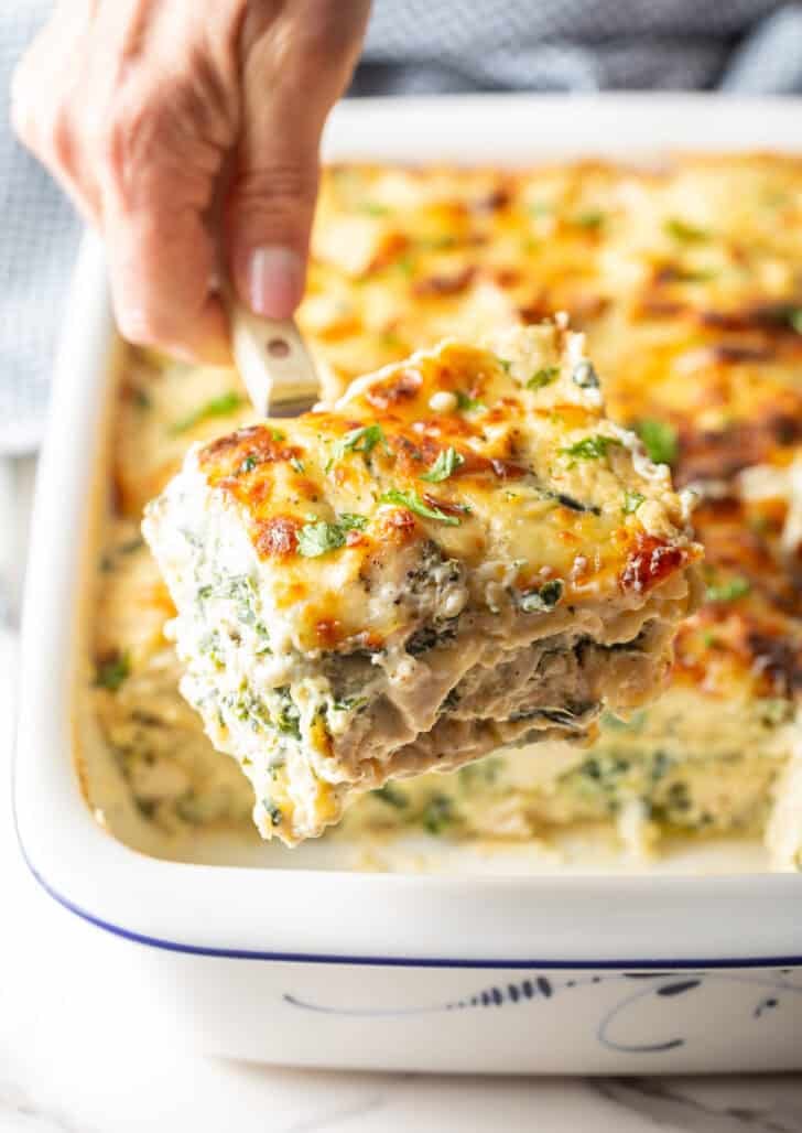 Hand using a serving spatula to lift a square of cheesy Alfredo chicken and spinach lasagna from a white baking pan.