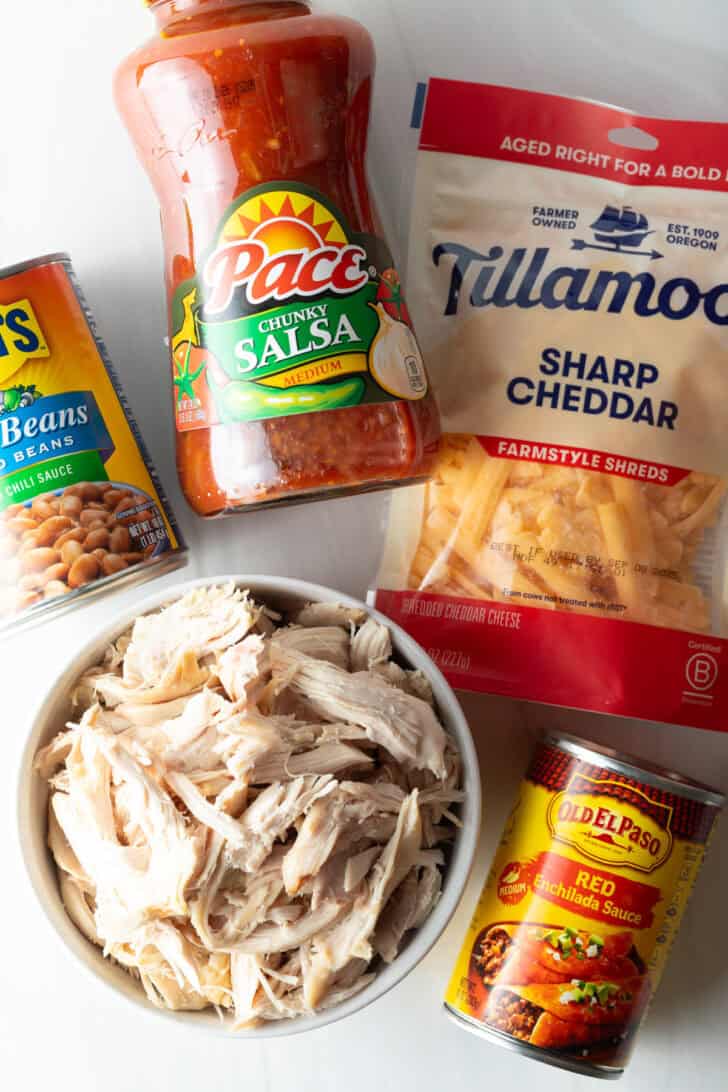 Top down view recipe ingredients: Bowl of shredded chicken, bottle of salsa, can of enchilada sauce, can of pinto beans and bag of shredded cheddar cheese.