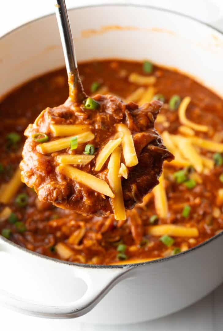 Ladleful of chicken chili and shredded cheese held to camera.