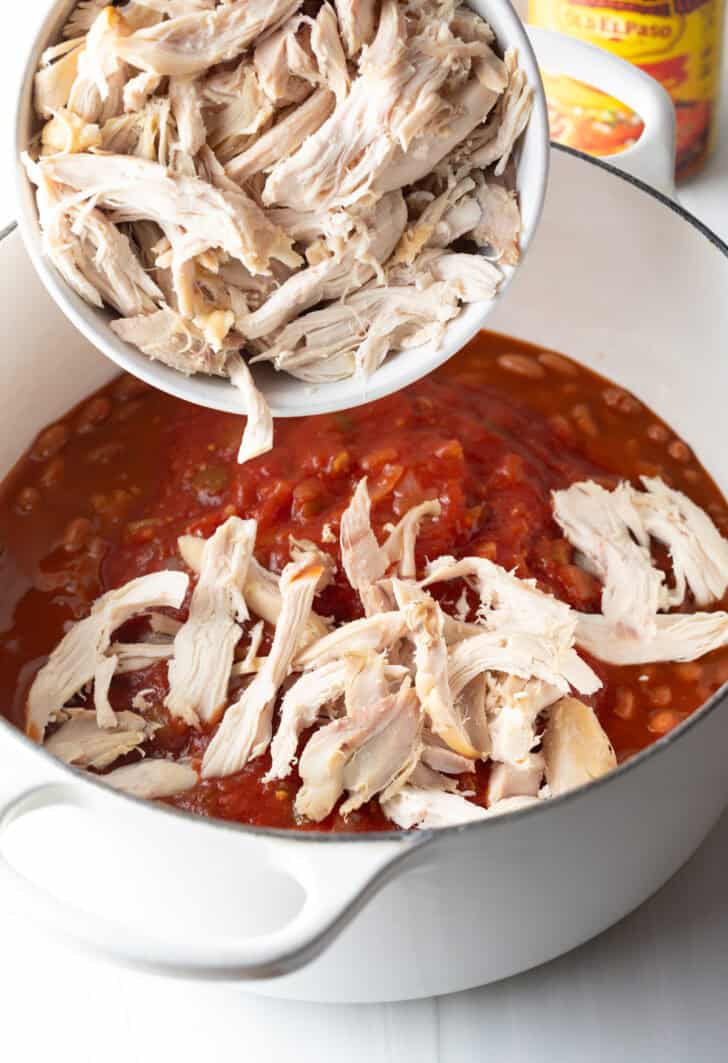Adding cooked shredded chicken to a pot of tomato sauce and beans.