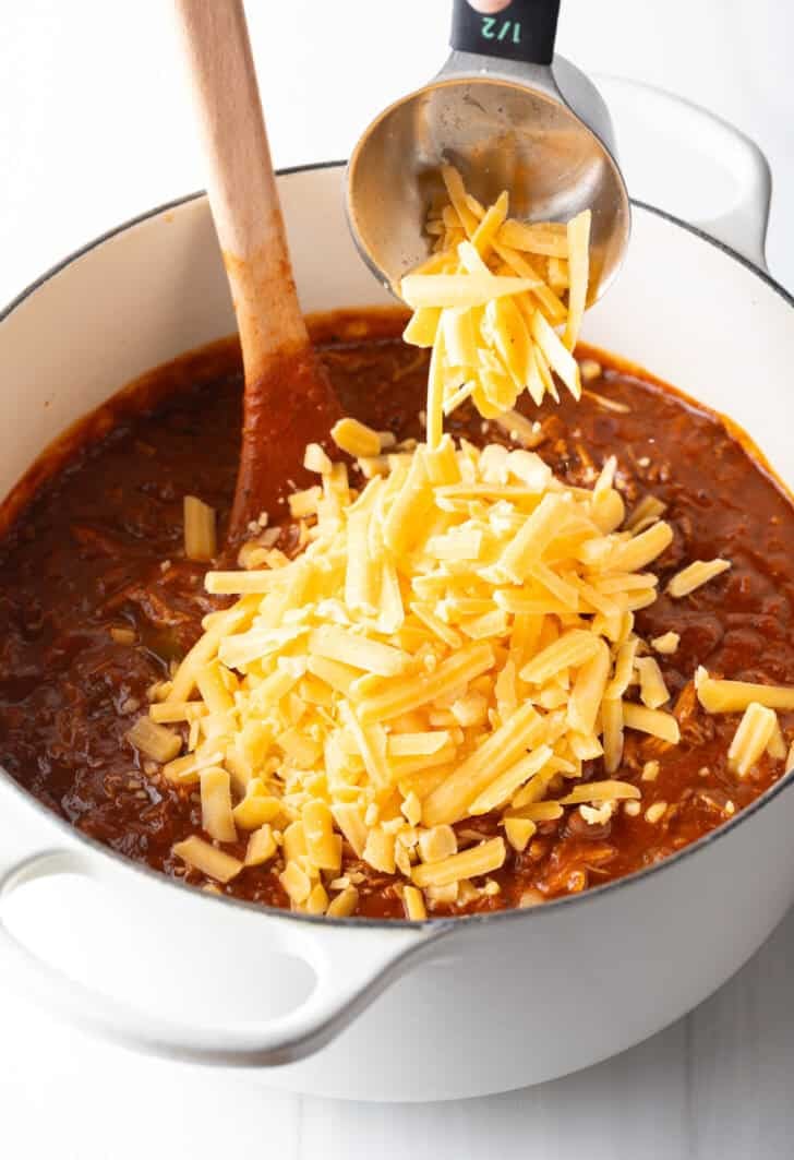 Adding shredded cheddar cheese to a large bot of chili.
