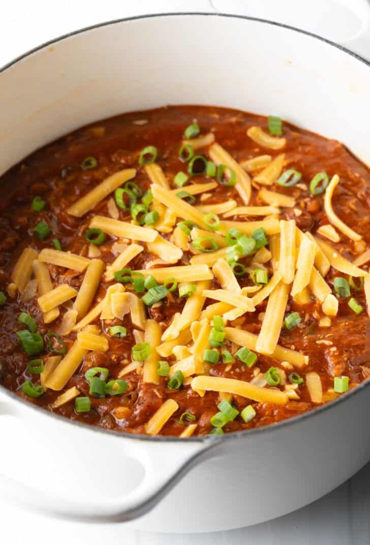 Large white soup pot with chicken chili, topped with chopped green onion and shredded cheddar cheese.