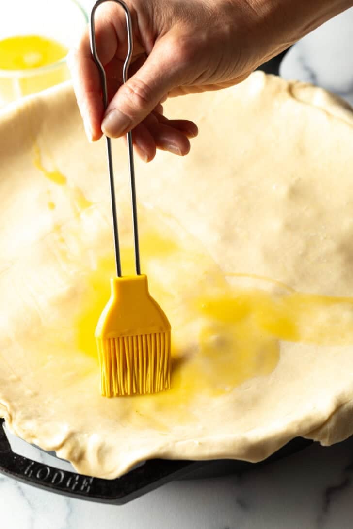 Hand using a yellow pastry brush to coat the top of puff pastry with melted butter.