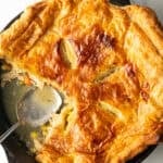 Skillet of chicken pot pie topped with golden brown puff pastry. A serving spoon has removed a quarter portion.