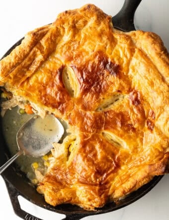 Skillet of chicken pot pie topped with golden brown puff pastry. A serving spoon has removed a quarter portion.