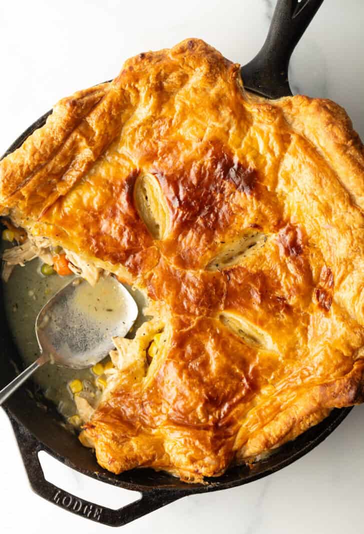 Skillet of chicken pot pie topped with golden brown puff pastry. A serving spoon has removed a quarter portion.