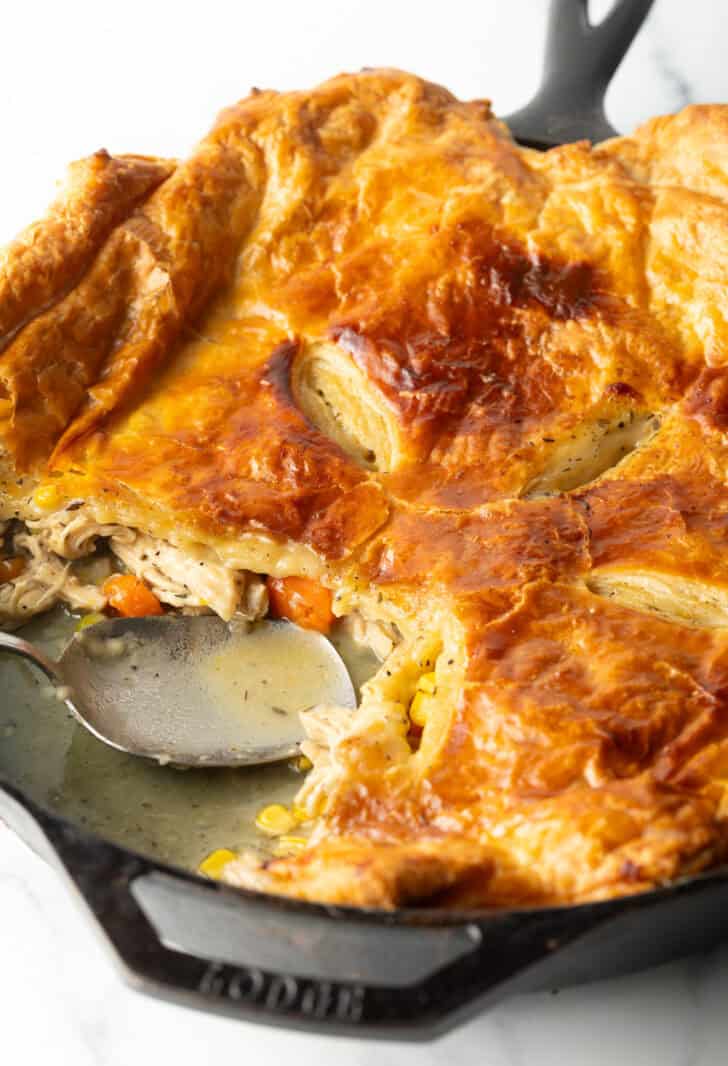 Skillet of chicken pot pie topped with golden brown puff pastry. A serving spoon has removed a quarter portion.