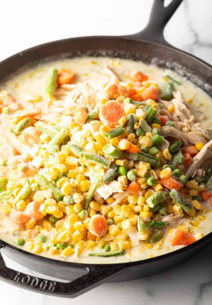 Adding frozen mixed vegetables to cast iron skillet with creamy gravy.